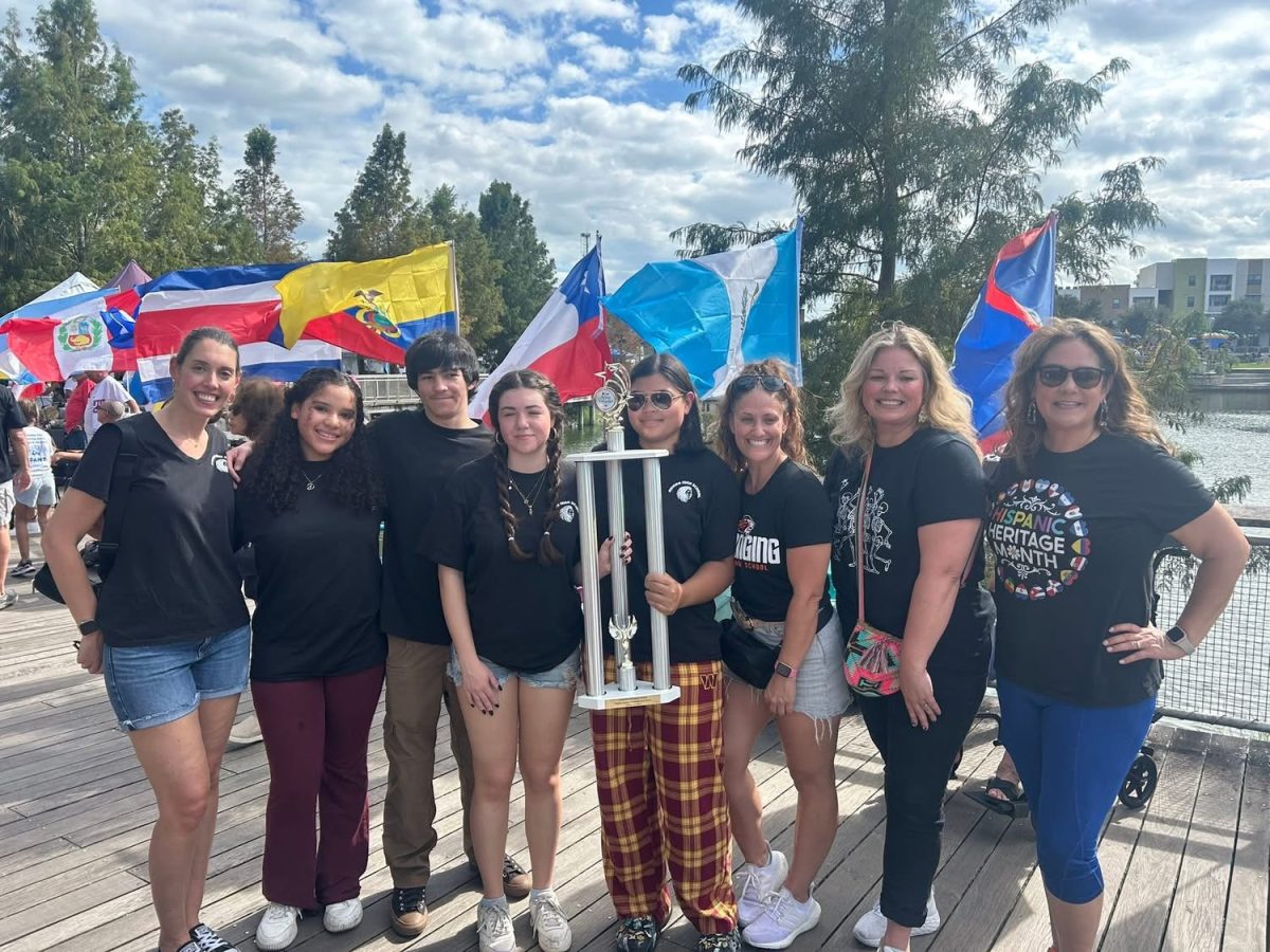Spanish Club members and sponsors (Ms. O’Connor, Hampton-Veloz, Cebreros, Galarreta, Osborne, Mrs. Charcap, Ms. MacCracken) pose with principal Dr. Daniel holding their winners trophy. They stand on the boardwalk of Center Lake Park, decorated with flags of several Spanish-speaking countries.