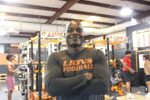 Sporting a Lions Football tee, Nordly Capi crosses his arms, posing inside the weight room during seventh period. He takes advantage of this time to further connect with and help his players and students.