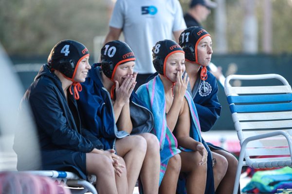Julia Cannaday, Tilly Glaser, Tessa Sims, and Zoe Belden watch the pool from afar. Some of their hands are to their mouths, spectating the excitement together, towels wrapped around them.